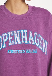 Fialová mikina se nápisy "COPENHAGEN" a "ATHLETICS SQUAD" v texturované bílé a modré barvě. Obsahuje kulatý výstřih a měkkou látku.