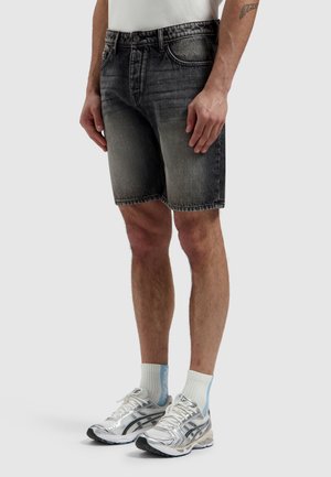 Man draagt vervaagde zwarte denim shorts, witte sportsokken en zilveren hardloopschoenen, staand tegen een effen lichte achtergrond.
