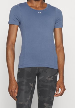 Person iført en tætsiddende kortærmet blå sportstrøje og grå/sort camouflage-mønstrede leggings, der står mod en ensfarvet baggrund.
