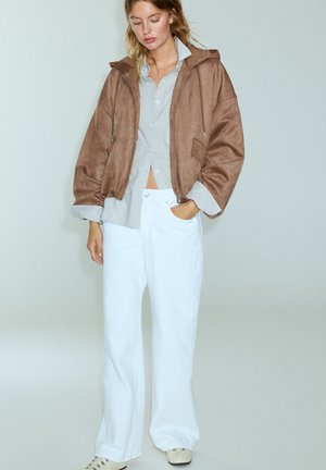 Femme portant une veste marron oversized, une chemise rayée bleue, un pantalon blanc à jambes larges et des chaussures claires, debout sur un fond uni.