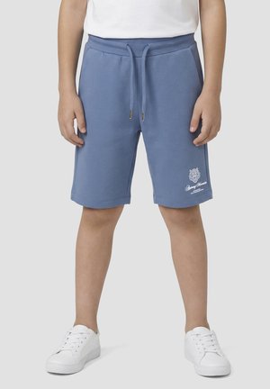 Bambino indossa pantaloncini blu con coulisse e tasche laterali e scarpe da ginnastica bianche, in piedi davanti a uno sfondo semplice.