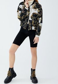 Veste noir et blanc avec motif chaîne dorée, assortie à un short noir déchiré et des bottes noires avec des lacets dorés et des semelles épaisses.