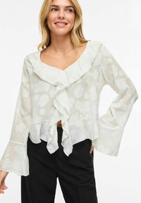Nasmejana ženska v belem cvetličnem prosojnem bluzonu z naborki spredaj in črnimi hlačami, ki pozira z eno roko na boku.