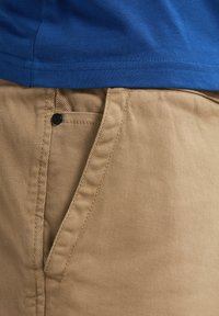 Beige bomuldsbukser med en struktureret overflade, der har en forlommem og sort knapdetalje, sammen med en blå t-shirt.
