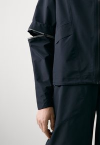 Navy waterproof jacket with a zippered arm pocket, clean lines, and a matte finish. Features adjustable cuffs and practical design elements.