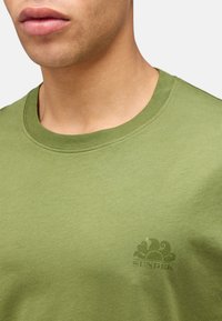 T-shirt in cotone verde oliva con scollo rotondo e maniche corte, caratterizzato da un logo tono su tono sul petto. Texture liscia.