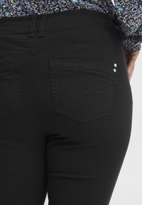 Svarta denimjeans med en slät textur, featuring två bakfickor med vinklade mönster och dekorativa metallaccenter.