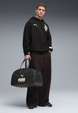 Mann trägt schwarzen Puma Hoodie und Hose, hält eine große schwarze Puma Sporttasche in seiner rechten Hand vor einem grauen Verlaufshintergrund.