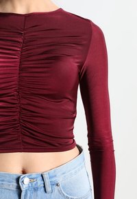Burgundská blůza s dlouhým rukávem a nařasenou texturou vpředu, s přiléhavým střihem, spárovaná se světle modrými džínami s vysokým pasem.