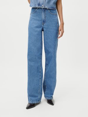 Kobieta ubrana w niebieskie jeansy z wysokim stanem i luźnym krojem, w połączeniu z czarnymi butami w szpic, na jednolitym tle.