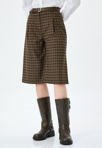 Pantaloni corti a quadri marroni, lunghi fino al ginocchio e con pieghe, dotati di una cintura marrone. Abbinati a stivali alti in pelle marrone scuro con dettagli con fibbia.