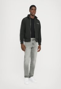 Sort zip-up bomberjakke med hvide striber, båret over en sort t-shirt. Lys grå jeans og hvide sneakers fuldender outfittet.