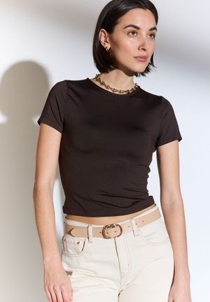 Frau mit kurzem dunklem Haar, die ein schwarzes, eng anliegendes T-Shirt, einen beigen Gürtel mit runder Schnalle, weiße Hose und eine goldene Halskette trägt und seitlich posiert.