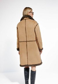 Beżowy płaszcz z owczej wełny z ciemnobrązowymi wykończeniami, z szerokim kołnierzem i kontrastowymi szwami. Długie rękawy i luźny krój.