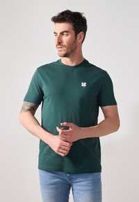 Groene katoenen T-shirt met korte mouwen en een ronde hals, voorzien van een klein wit klaverlogo aan de linkerzijde. Soepele textuur.