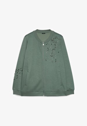 Veste verte à fermeture éclair avec col et poignets côtelés, décorée d'embellissements en perles noires et blanches éparpillés sur la poitrine et la manche gauche.