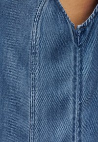 Κοντινή λήψη υφάσματος από τζιν με ανοιχτό μπλε χρώμα, εμφανή υφή και λεπτομέρεια φερμουάρ στη λαιμόκοψη με επάνω ραφή.