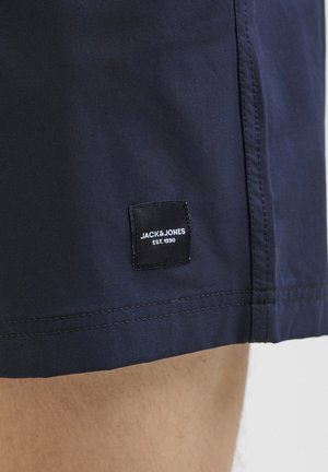 Marineblaue Shorts aus glattem Stoff, mit einem schwarzen Etikett mit weißem Text "JACK & JONES EST. 1990", das an der Seite aufgenäht ist.