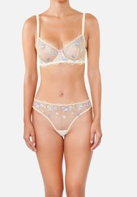 Femme portant un ensemble de lingerie beige transparent avec des broderies florales bleues et roses, posant devant un fond clair.