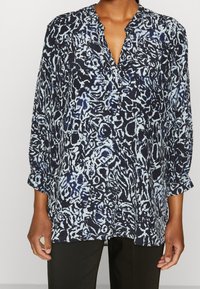 Mujer vistiendo una blusa de manga larga de corte holgado con un patrón abstracto en azul y negro y un escote en V, combinada con pantalones negros.