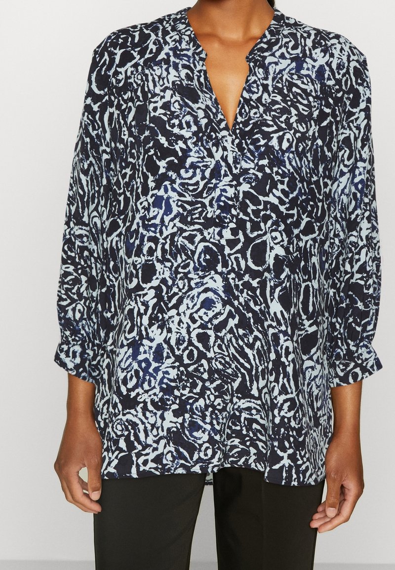 Mujer vistiendo una blusa de manga larga de corte holgado con un patrón abstracto en azul y negro y un escote en V, combinada con pantalones negros.