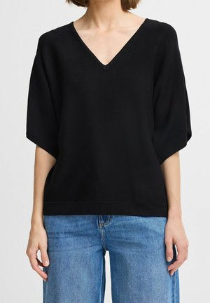 Frau trägt ein schwarzes, locker sitzendes Top mit V-Ausschnitt und kurzen Ärmeln, kombiniert mit hellblauen Jeans, vor neutralem Hintergrund.