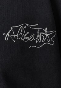 Tessuto nero con un logo bianco scritto a mano, "Allsaints", visualizzato in uno stile corsivo fluido con variazioni nello spessore delle linee.