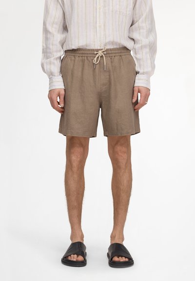 Homme portant un short beige à cordon, une chemise à manches longues rayée claire et des sandales noires à enfiler, debout devant un fond blanc.
