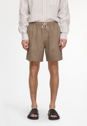 Mężczyzna ubrany w beżowe szorty ze ściągaczem, jasną pasiastą koszulę z długim rękawem i czarne klapki, stojący na białym tle.