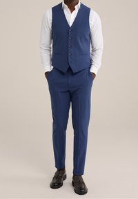 Námořnická modrá vesta s pěti knoflíky, pod ní bílá košile a ladící kalhoty. Má slim fit střih a strukturovanou tkaninu. Temná obuv doplňuje celý vzhled.