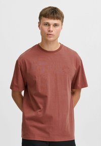 Rustfarvet bomulds t-shirt med korte ærmer, rund halsudskæring og subtilt cirkulært logo på brystet. Glat tekstur og afslappet pasform.