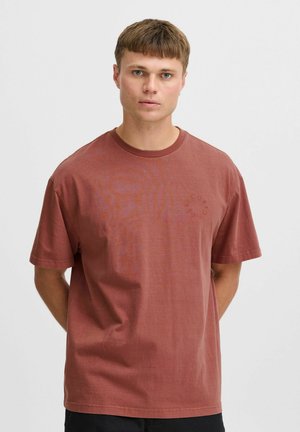 Ceglastoczerwony bawełniany t-shirt z krótkimi rękawami, okrągłym dekoltem i subtelnym okrągłym logo na piersi. Gładka tekstura i luźny krój.