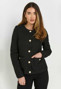 Musta teksturoitu takki, jossa on pyöreä kaula-aukko, kultaiset napit ja kaksi etutaskua. Kankaassa on hieno kiilto ja se on muotoiltu istuvaksi.