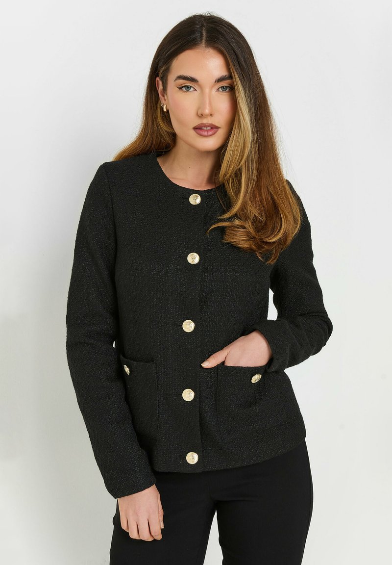 Musta teksturoitu takki, jossa on pyöreä kaula-aukko, kultaiset napit ja kaksi etutaskua. Kankaassa on hieno kiilto ja se on muotoiltu istuvaksi.