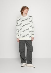 White knit sweater with black 'CARLO COLUCCI' text pattern, ribbed crew neck, and cuffs. Paired with gray cargo pants and canvas sneakers.
