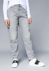 Γκρι τζην παντελόνι με χαλαρή γραμμή, ψηλή μέση και κλασικό σχέδιο πέντε τσέπες, σε συνδυασμό με λευκά αθλητικά παπούτσια με μαύρες λεπτομέρειες.