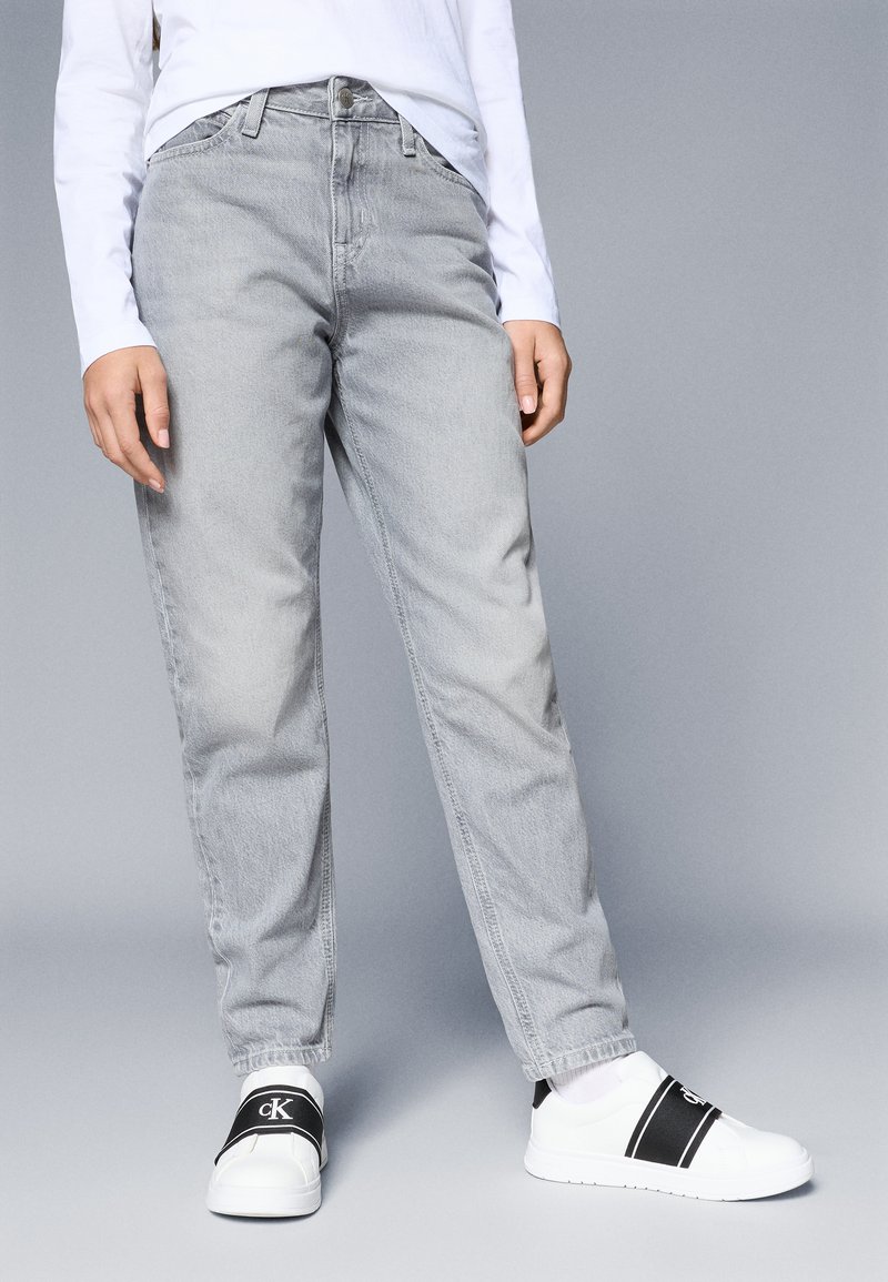 Γκρι τζην παντελόνι με χαλαρή γραμμή, ψηλή μέση και κλασικό σχέδιο πέντε τσέπες, σε συνδυασμό με λευκά αθλητικά παπούτσια με μαύρες λεπτομέρειες.