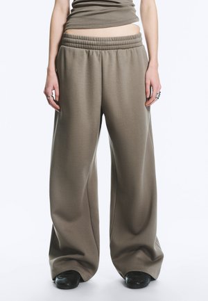 Osoba ubrana w luźne, szerokie spodnie dresowe w kolorze taupe z elastycznym paskiem w talii, zestawione z czarnymi butami i dopasowanym krótki topem.