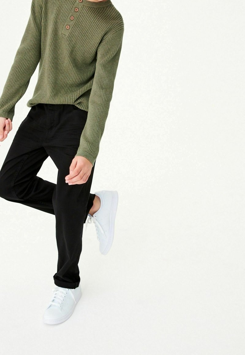 Personne en pull tricoté vert olive, pantalon noir et baskets blanches, levant une jambe pliée au niveau du genou sur un fond uni.