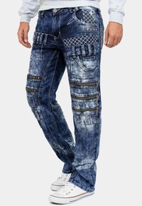 Pantaloni cargo in denim blu scuro con finitura sbiadita, numerose tasche con zip e dettagli cuciti. Presenta motivi decorativi e accessori metallici.