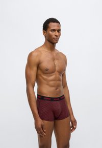 Cuecas boxer vermelhas com padrão e cós preto com o logótipo "HUGO". O design inclui formas geométricas e um ajuste justo.