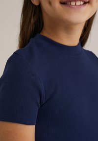 Tmavě modrý žebrovaný top s krátkým rukávem a vysokým výstřihem. Materiál vypadá měkký a strukturovaný, blízko přiléhající k tělu.