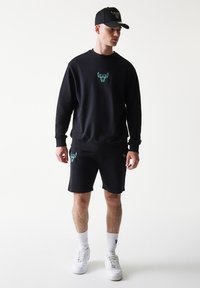 Μαύρο βαμβακερό φούτερ και σορτς με τουρκουάζ τυποποιημένα λογότυπα ταύρου. Συνδυασμένα με μαύρο καπέλο που φέρει παρόμοιο λογότυπο. Λευκά αθλητικά παπούτσια και κάλτσες.