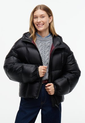 Junge Frau lächelt, trägt eine schwarze Steppjacke über einem grauen Zopfpullover und blauen Jeans, die Hände justieren den Reißverschluss der Jacke.