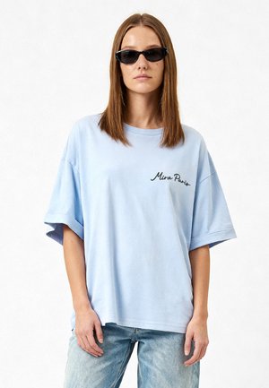 Femme aux cheveux bruns et lisses, portant des lunettes de soleil noires, un t-shirt ample bleu clair avec le texte « Mira Paris » et un jean bleu clair.