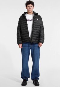 Fekete steppelt dzseki cipzáros elülső résszel, kapucnival és steppelt mintával, fehér inggel és kék, bő szárú farmerral párosítva. Fekete lábbeli.