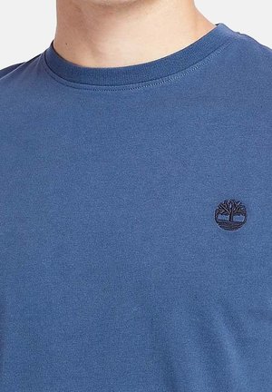 Homme portant un t-shirt bleu à col rond avec un petit logo brodé en forme d'arbre noir sur le côté gauche de la poitrine.