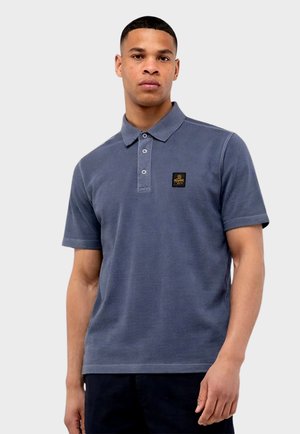 Giovane uomo che indossa una polo blu a maniche corte con una toppa con logo sul petto a sinistra, in piedi davanti a uno sfondo chiaro e semplice.
