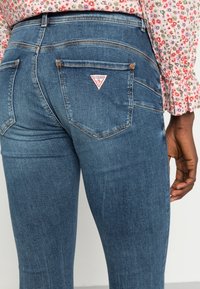 Jeans de mezclilla azul con un corte ajustado, bolsillos traseros con un parche de logo triangular y un sutil desgaste en la tela para darle textura.