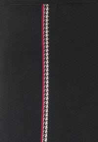 Černá látka s texturovaným povrchem, s vertikálním červeným pruhem a houndstooth vzorem v bílé a černé barvě lemujícím pruh.